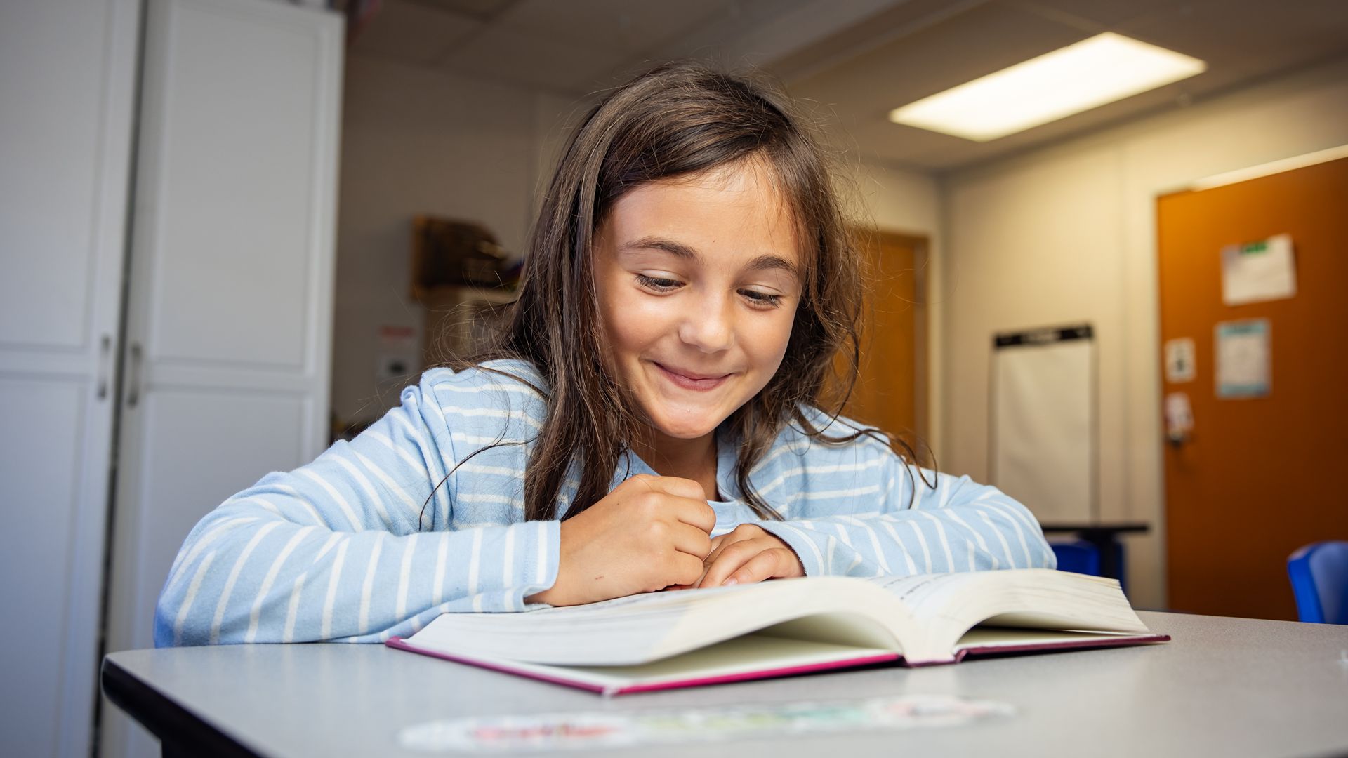 Smiling young girl reading a book at a classroom desk