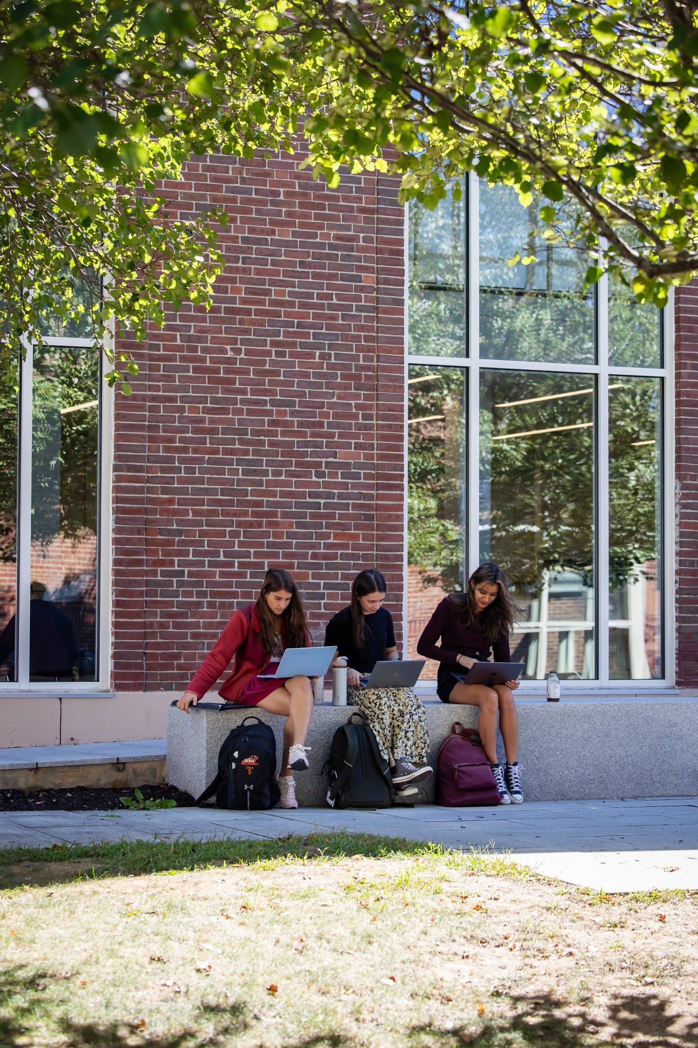 Three students sitting outside a brick school building working on laptops